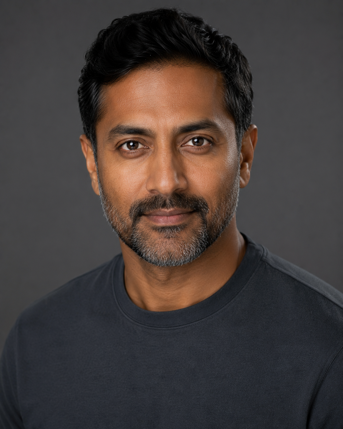 Real headshot the site owns. Controlled studio light, soft key from upper left, clean seamless mid-grey background, visible round catchlight at roughly 10 o’clock. Shot at typical headshot crop (top of head just inside frame, bottom around collarbone). Must be genuinely good — this is not a straw man.