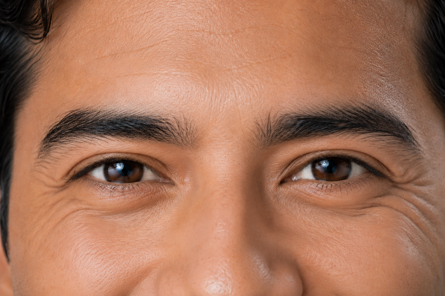 The SAME actor, same crop, same light, same session, now producing a genuine enjoyment smile driven by an actual thought. Lower eyelid visibly raised, cheeks up, skin bunched below the eye, crow’s feet at the outer corner, brow marginally lowered. Mouth still outside the frame. The two crops must be geometrically identical so the reader’s eye can only compare the eye region.