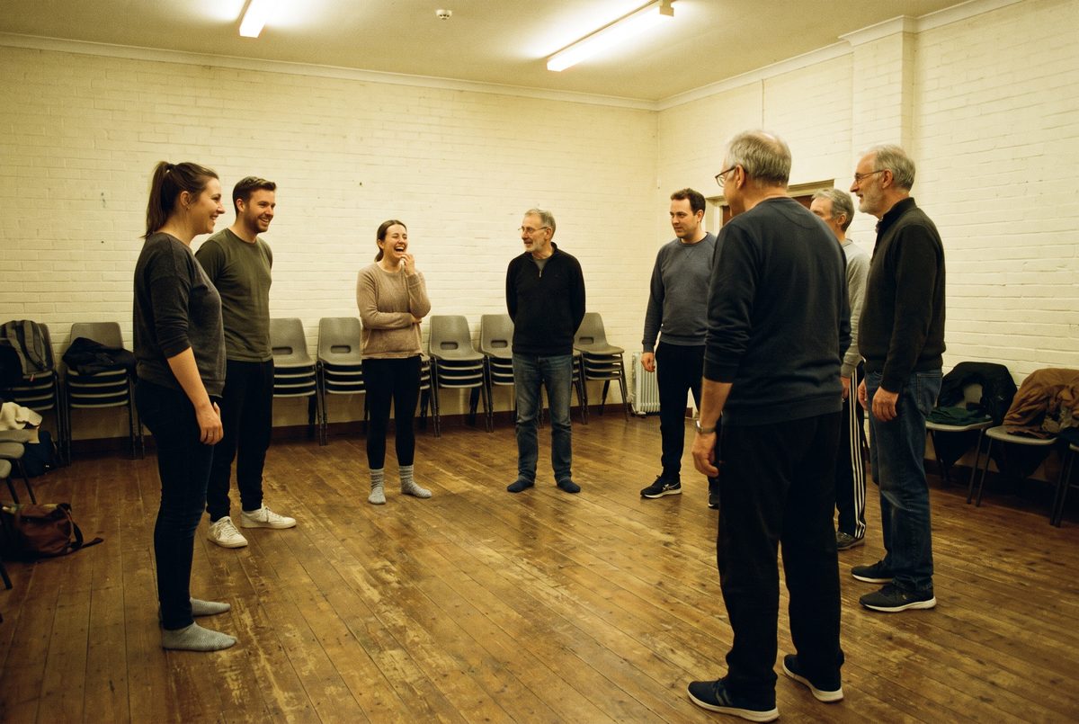 A mixed-age adult acting class standing in a loose circle in a rehearsal room