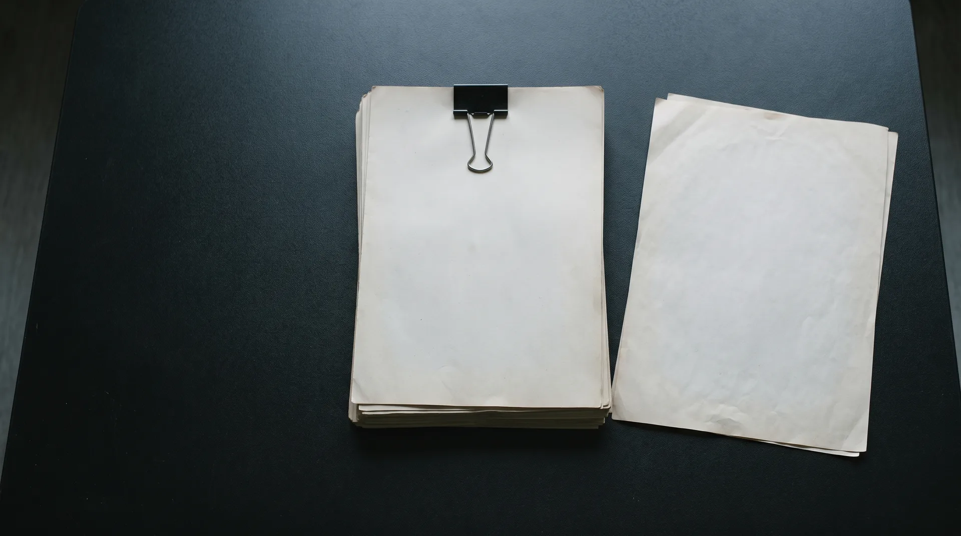 A thick clipped document and a single loose sheet lying side by side on a dark table