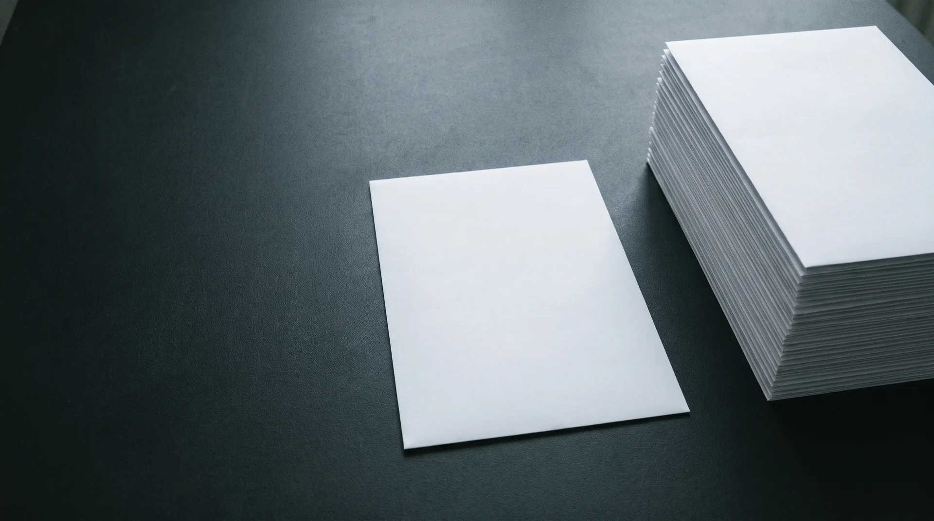 A single sheet of paper on a dark table beside a tall stack of many identical sheets