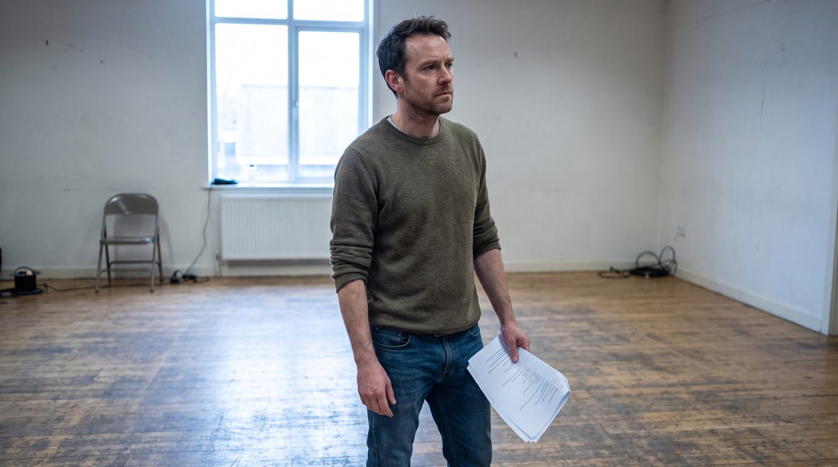 An actor in a bare rehearsal room holding script pages, attention fixed on someone off camera