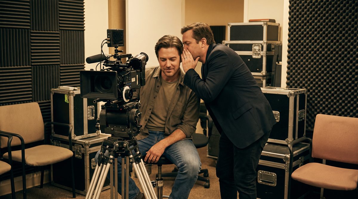 A director leaning in to give a quiet note to an actor beside a camera in a casting room