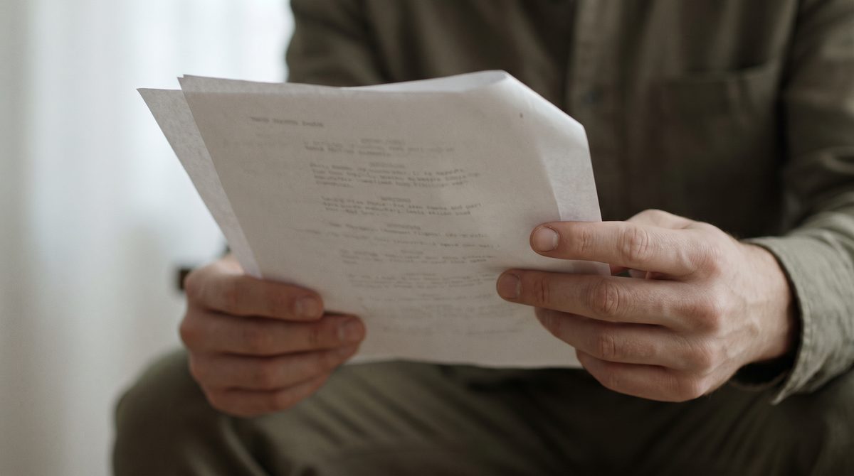 Hands holding loose script pages at chest height, thumb marking the place, one corner folded