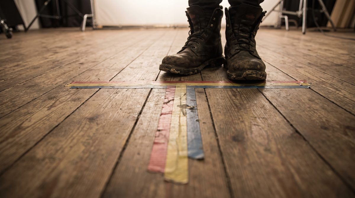Close view of gaffer tape marks forming a T on a studio floor beside an actor's feet