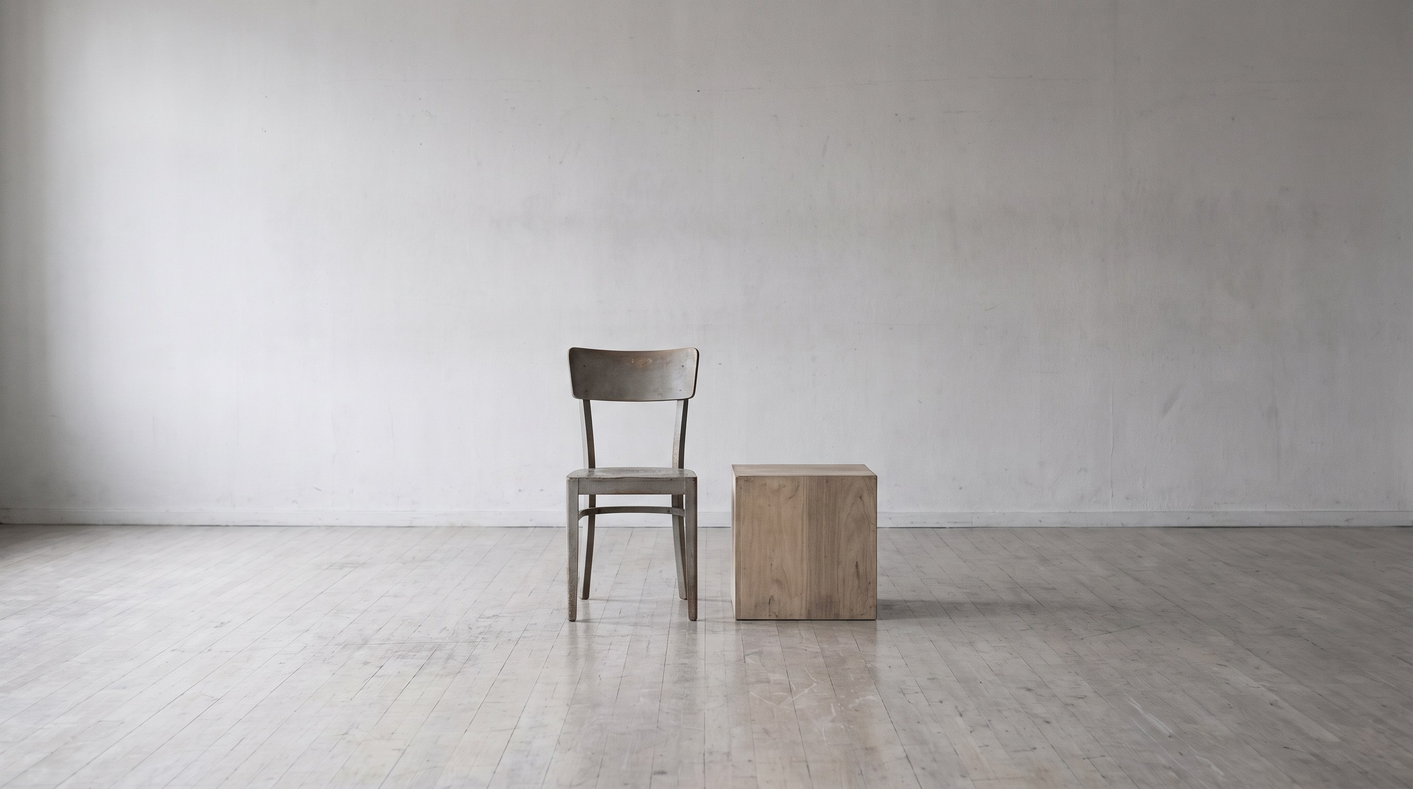 A plain wooden chair and a simple wooden cube standing on a bare floor in an empty rehearsal room