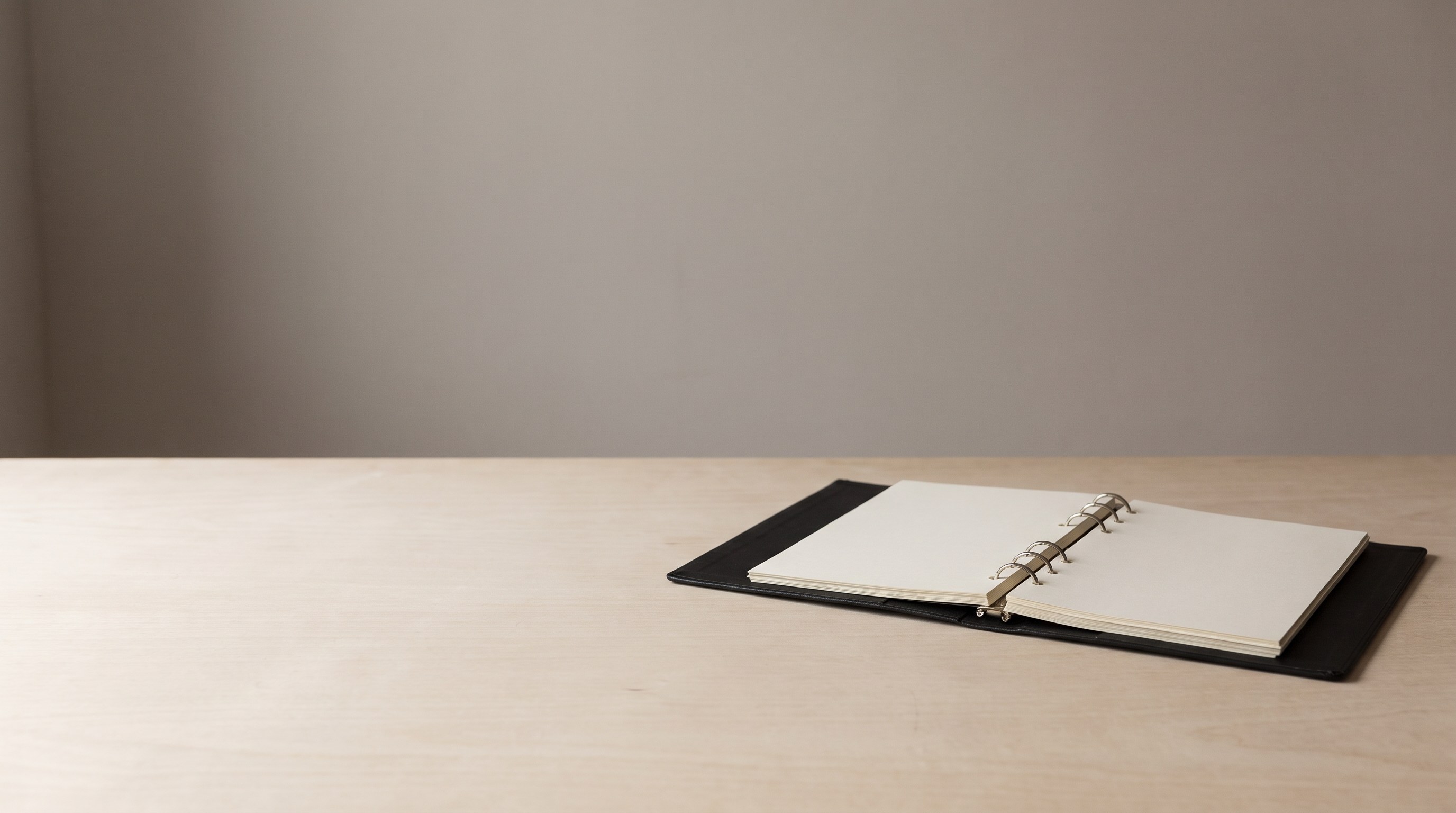 A slim black ring binder lying open on a plain wooden table, its pages blank