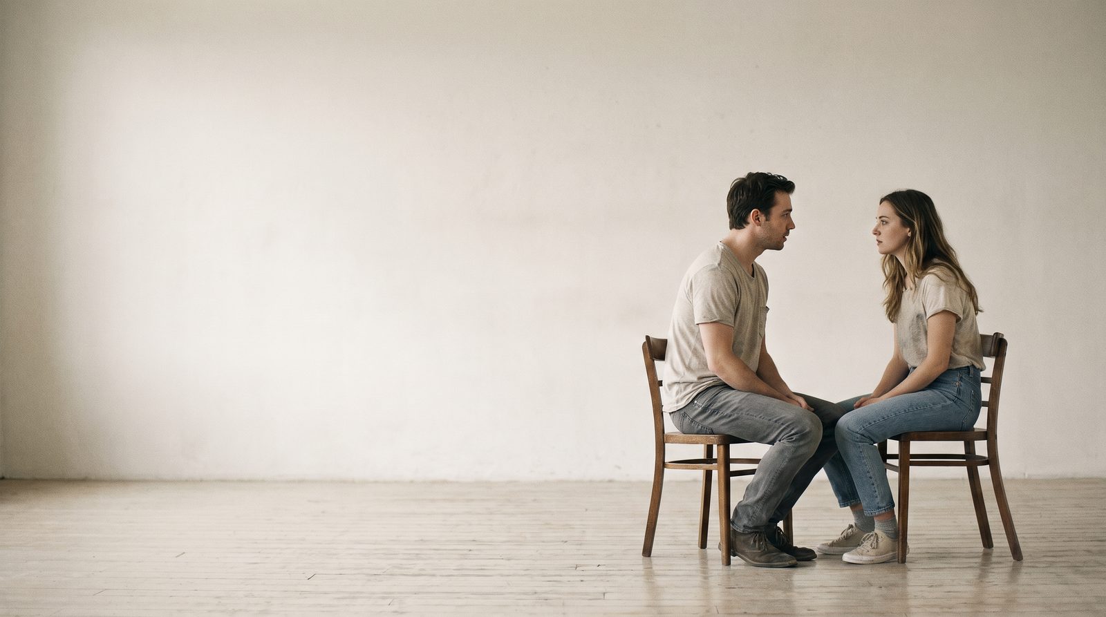 Two actors sitting knee to knee on plain chairs looking directly at each other in a rehearsal studio