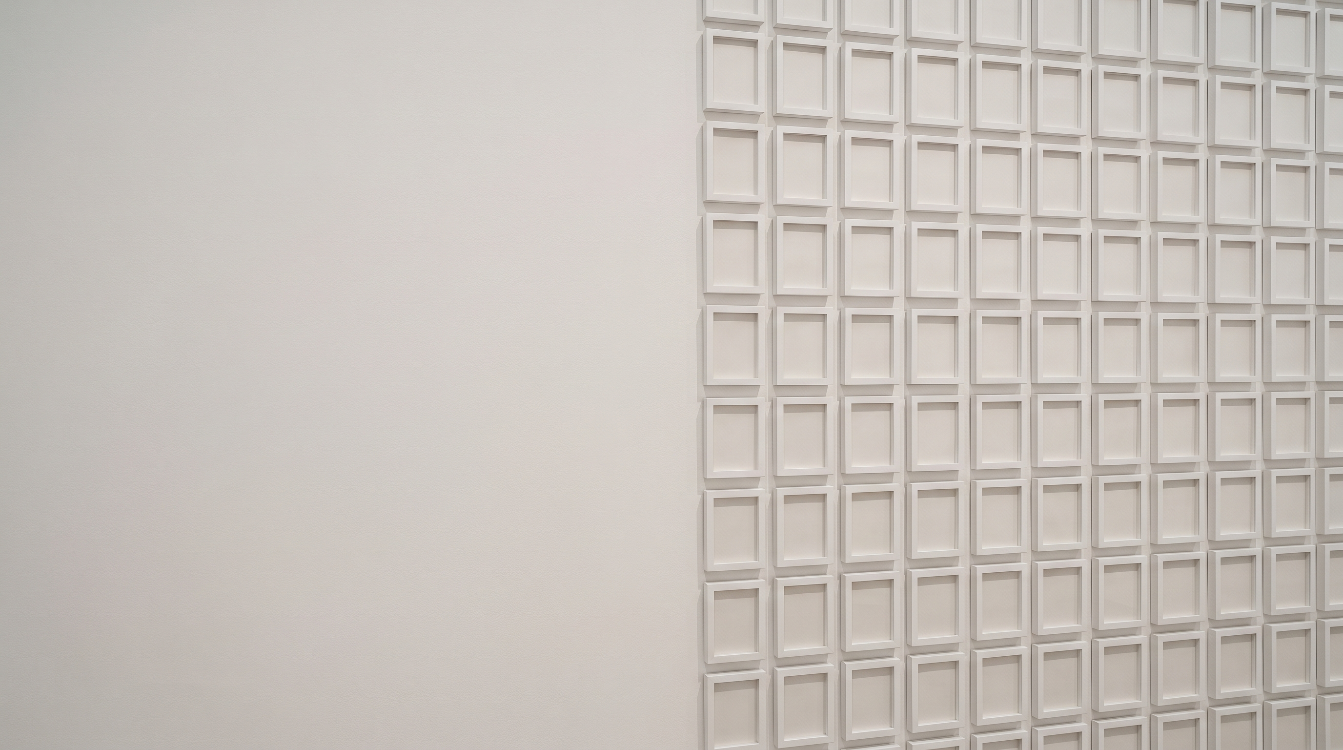 A dense grid of small empty white picture frames hung in rows on a plain wall