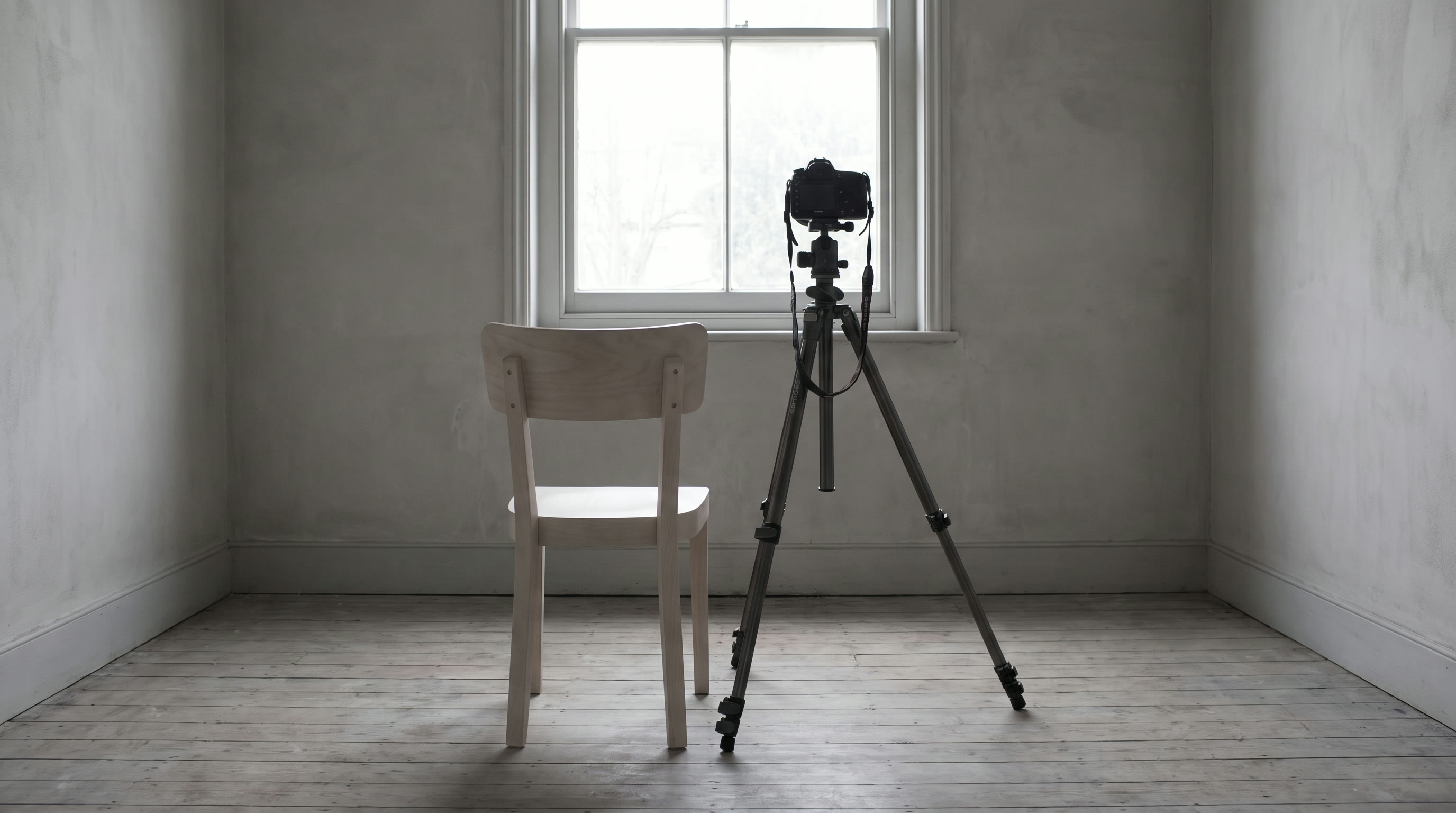 An empty plain chair positioned close beside a camera tripod at eye height in a quiet room