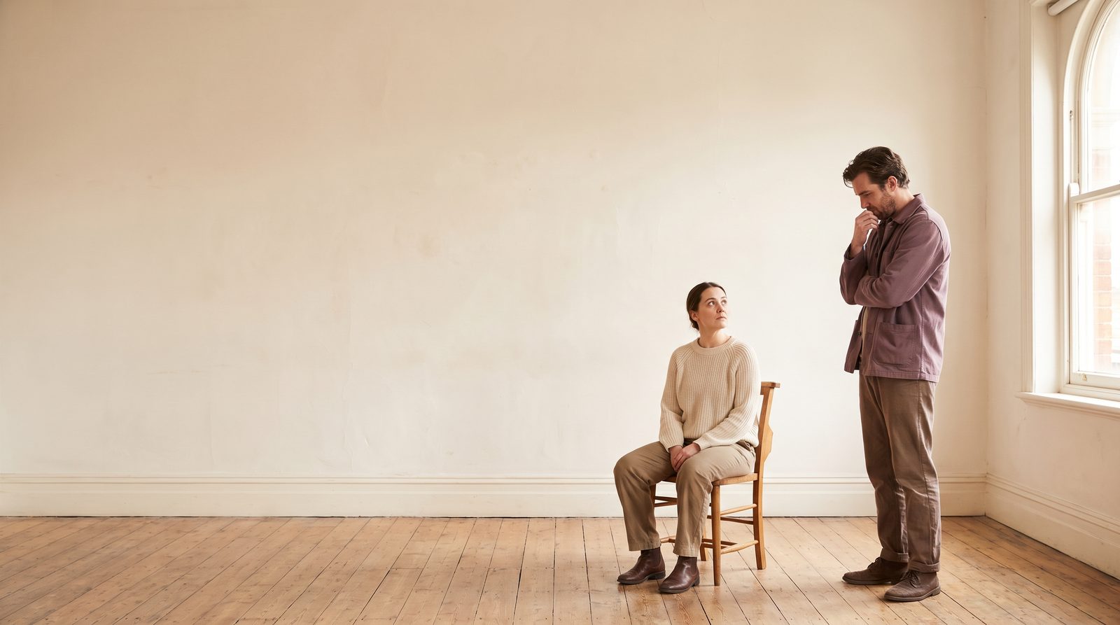 Two actors in a scene, one standing and one seated, watching each other