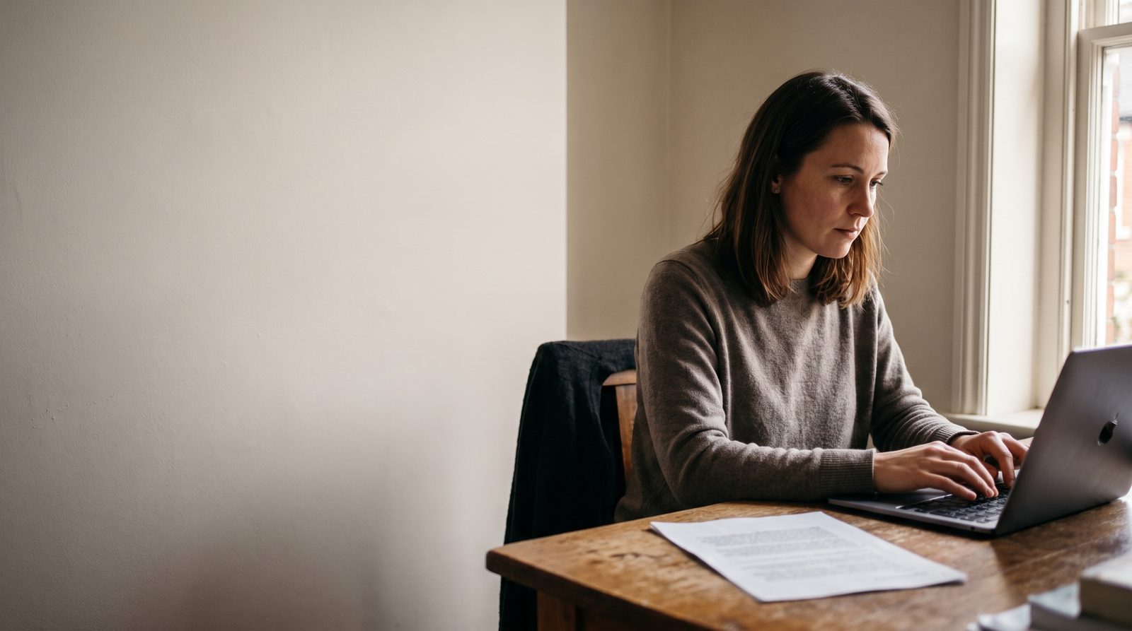 An actor typing at a laptop with a printed page on the desk beside it