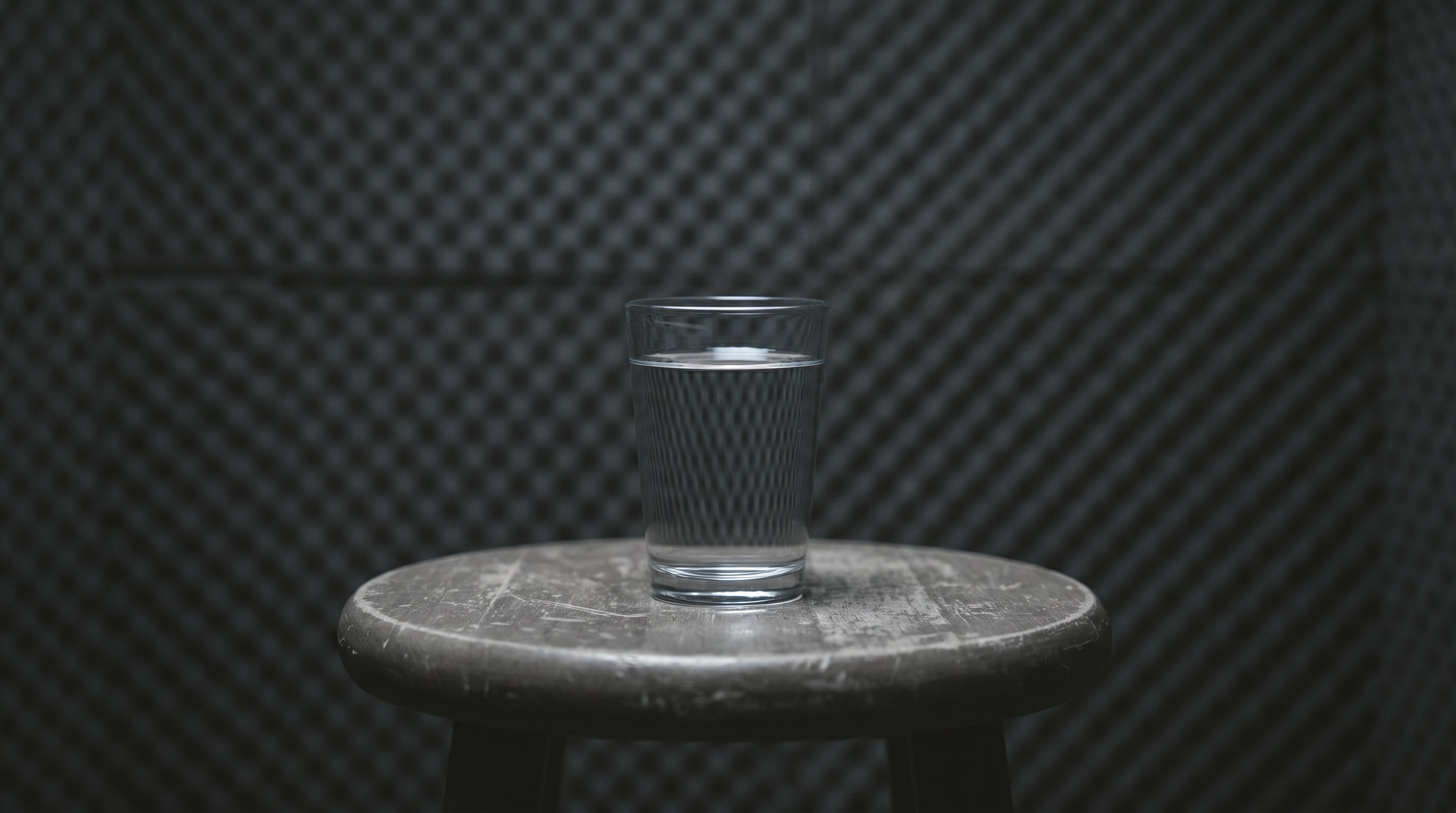 A plain glass of water on a wooden stool in a dim recording booth