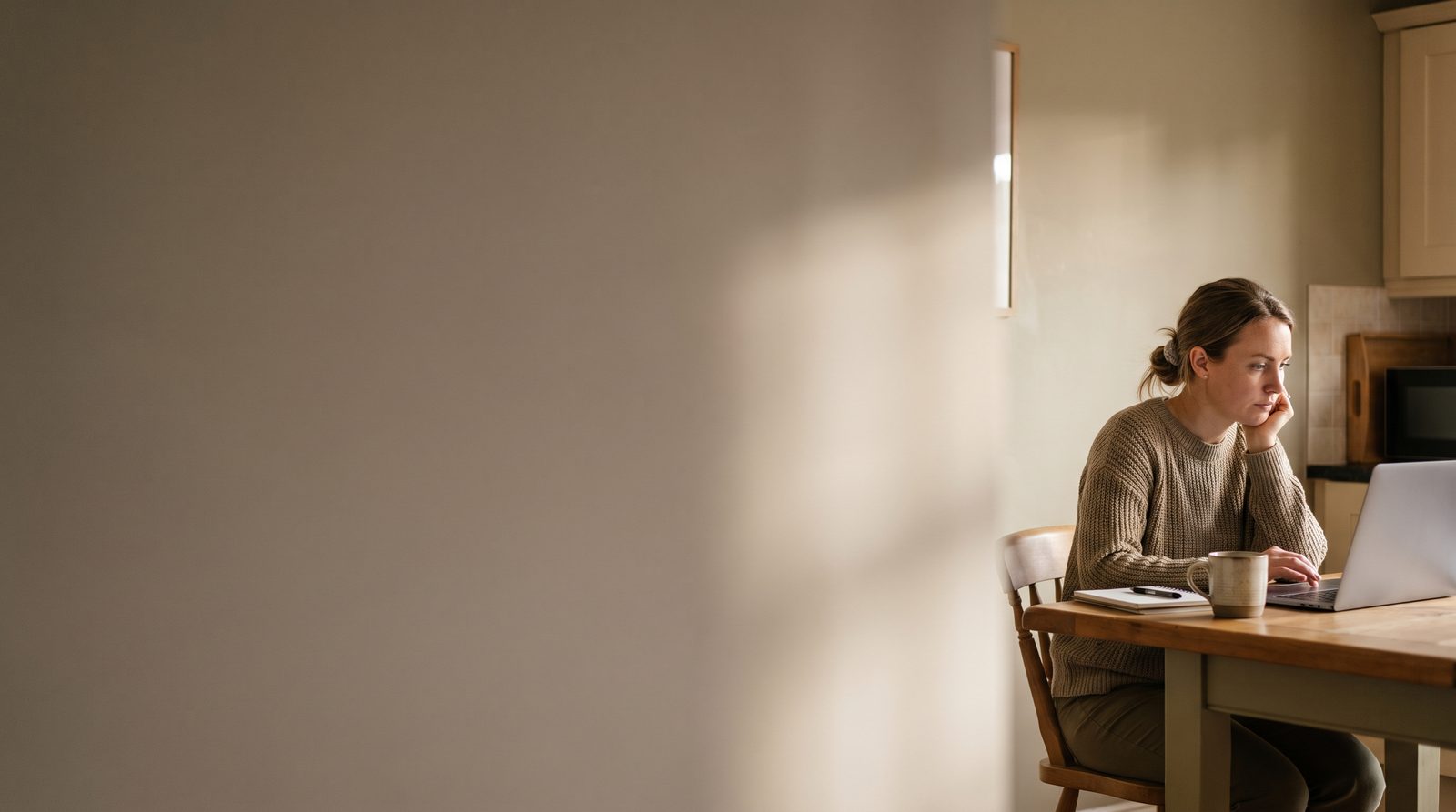 An actor at a kitchen table looking at a laptop, sunlight across a plain wall behind