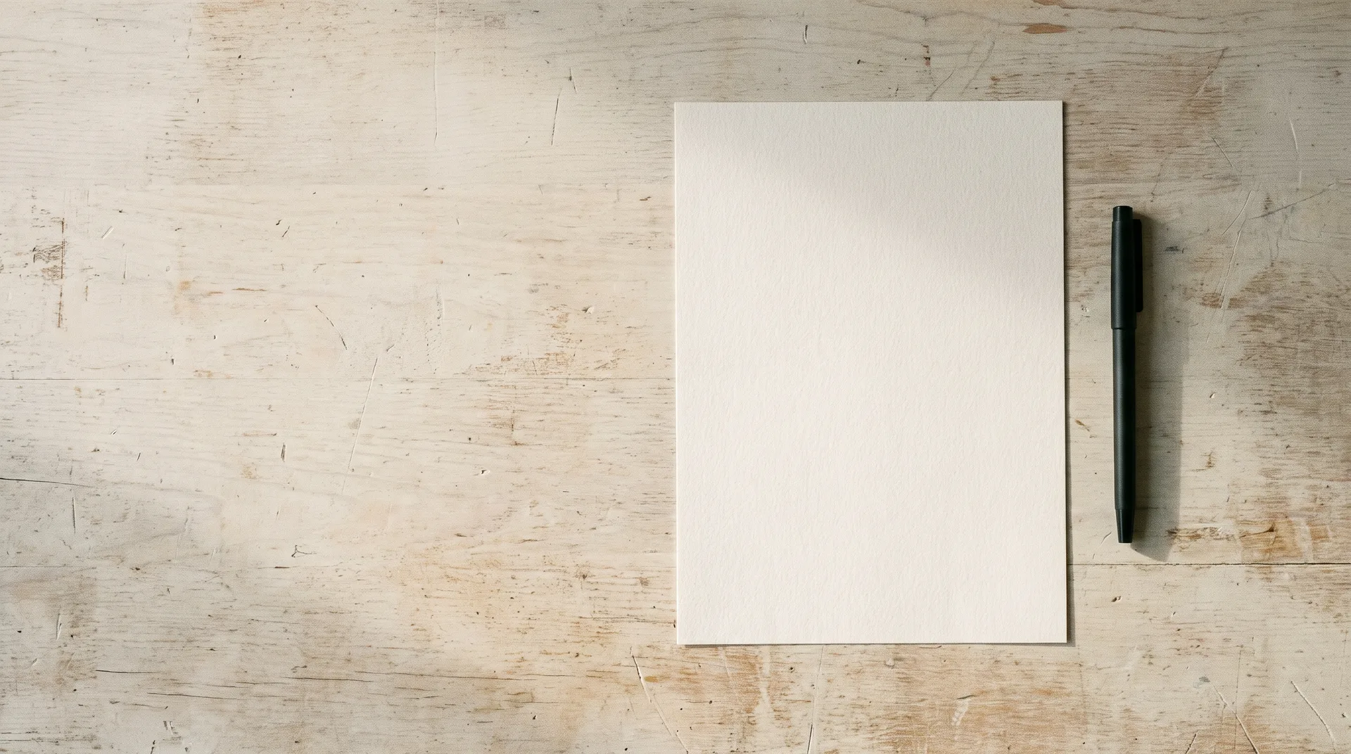A single blank sheet of paper and a pen on a pale wooden desk