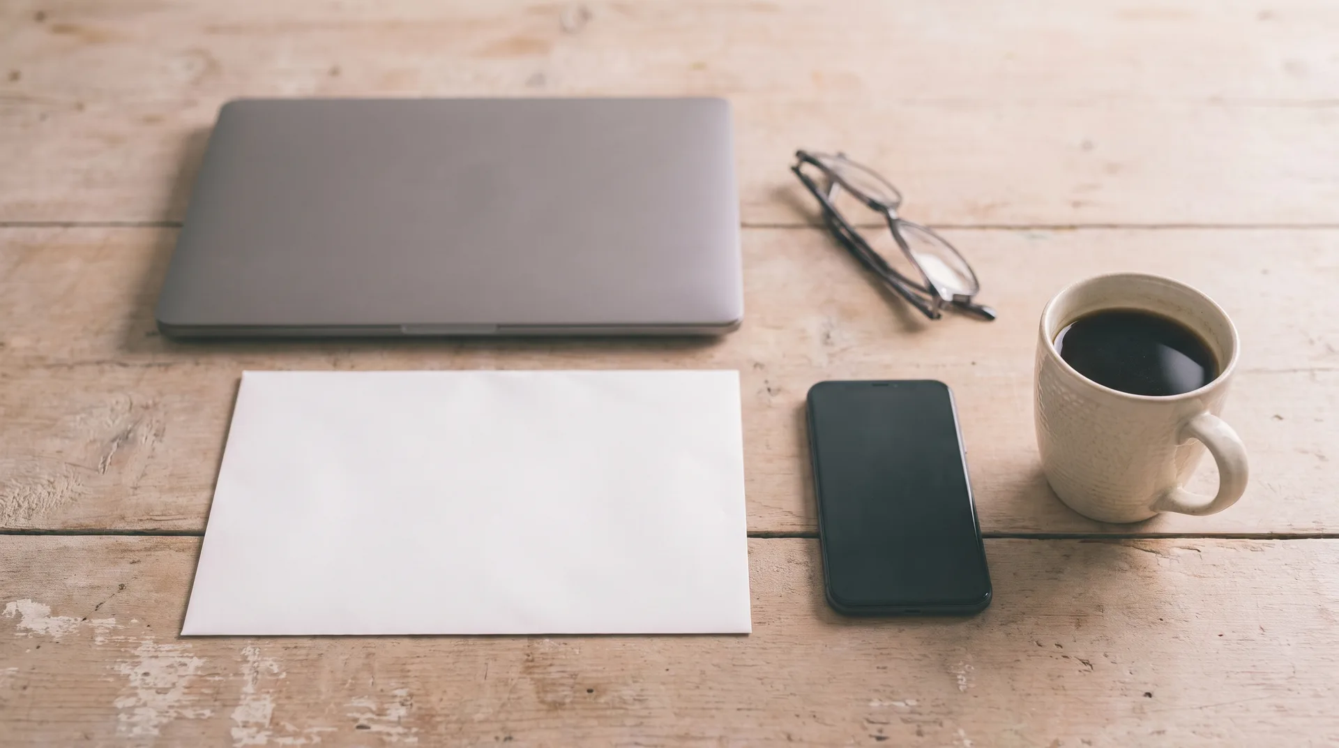A closed laptop, a blank white envelope, reading glasses and a coffee cup on a bare wooden desk