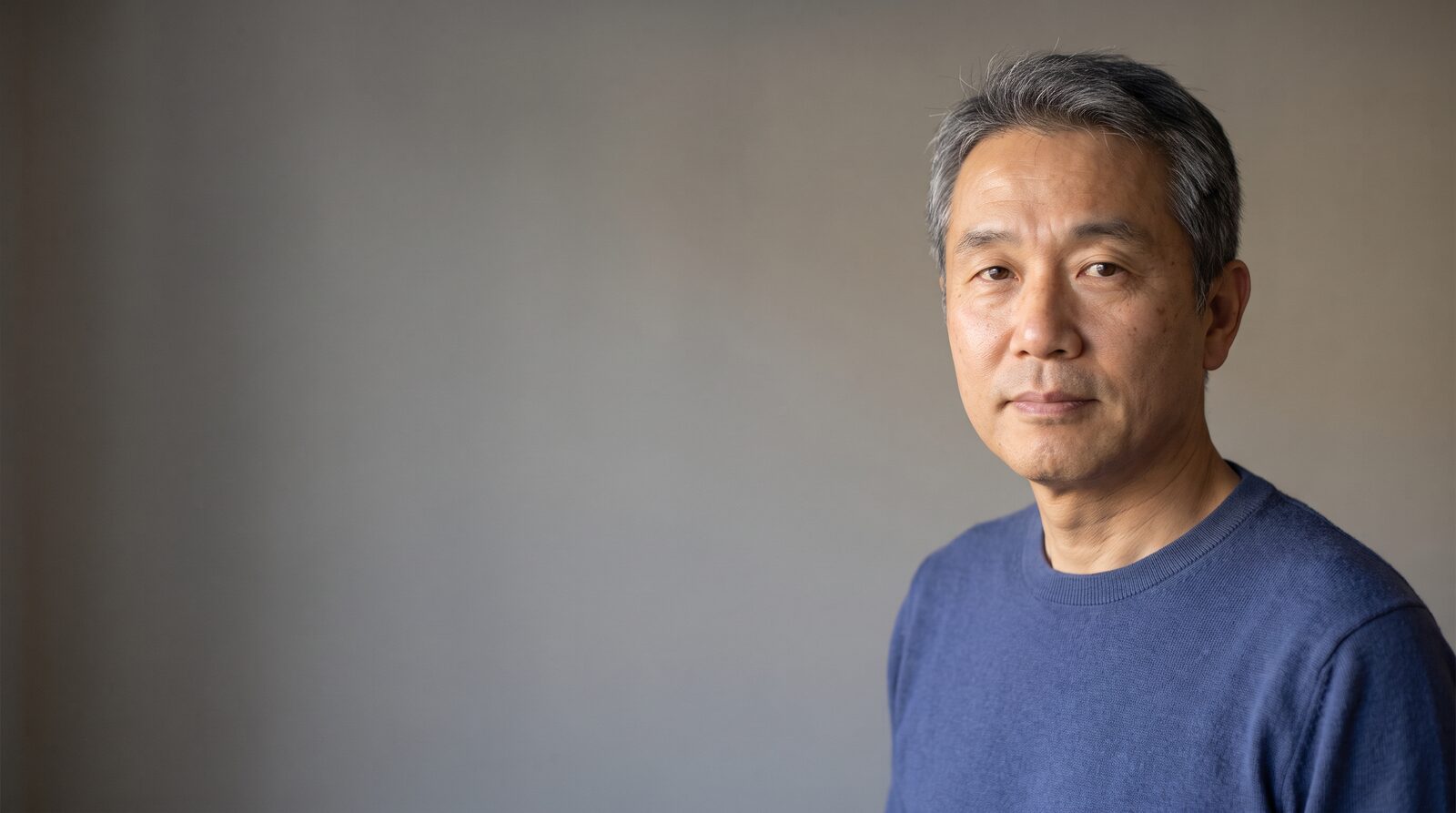 An actor framed chest-up against a plain mid-grey wall, clearly separated from the background
