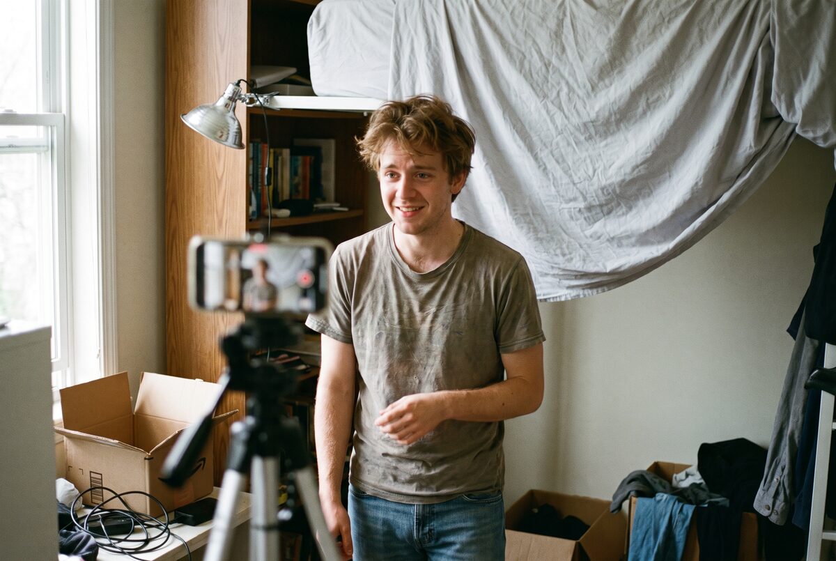 A young actor standing in a bedroom set up for filming, with a phone on a tripod and a plain backdrop